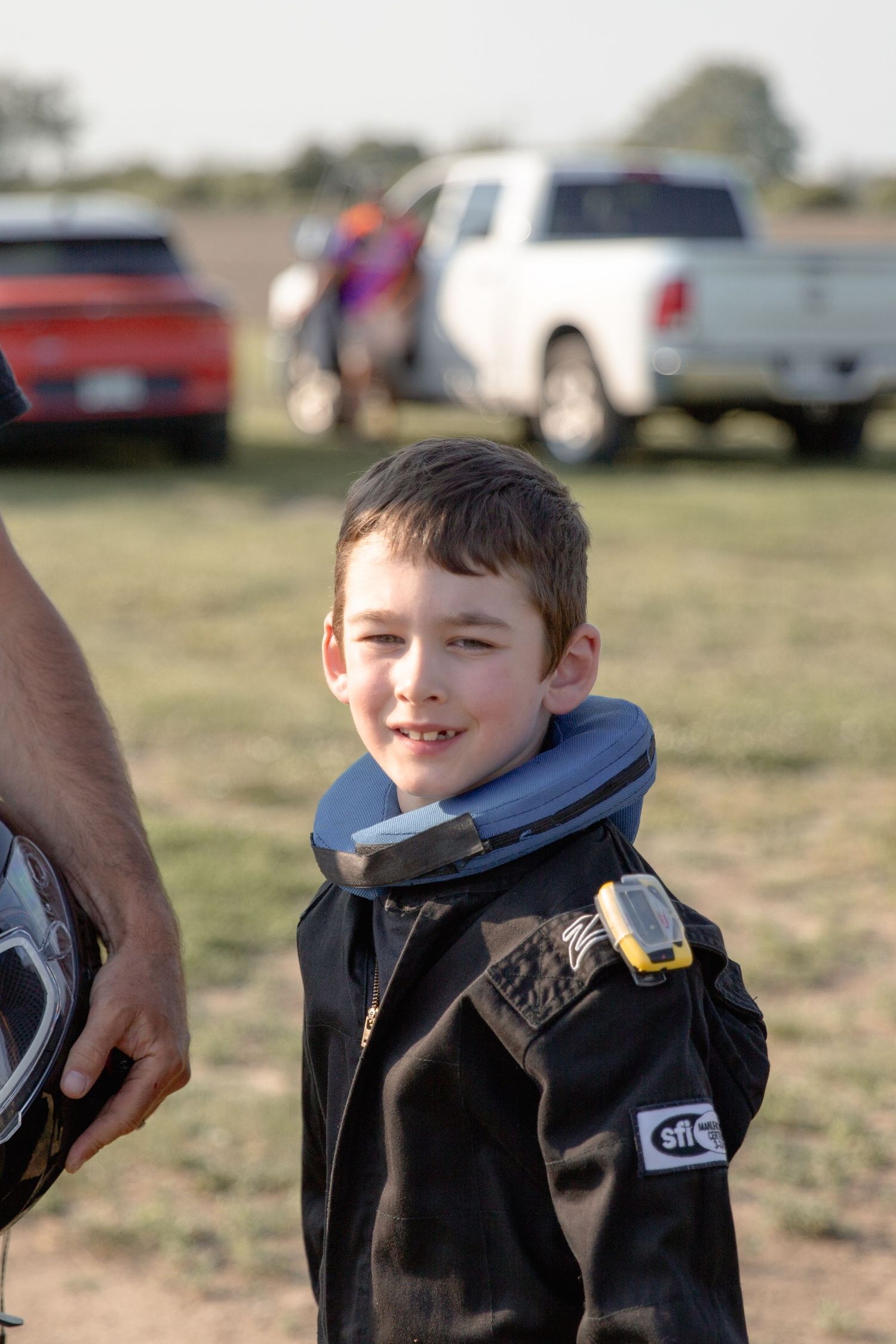 A young child in a blue hoodie and black jacket smiles while outdoors with cars visible in the background.