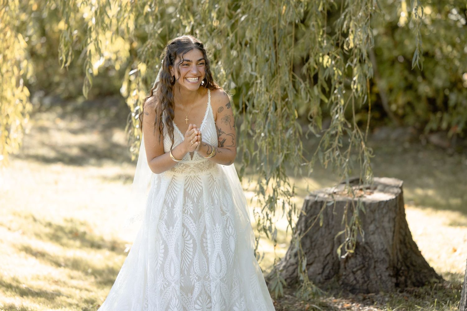 A person in a flowing white lace wedding dress stands in a sunlit garden setting under trees.