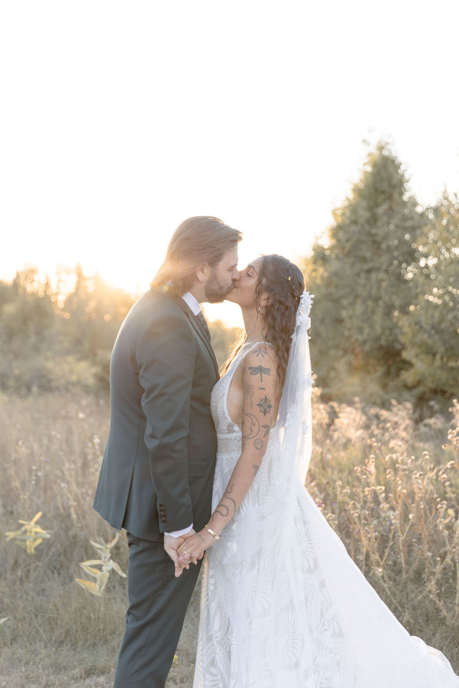 A romantic wedding couple shares a tender kiss in a sunlit field at golden hour as her white lace dress and veil flow softly.
