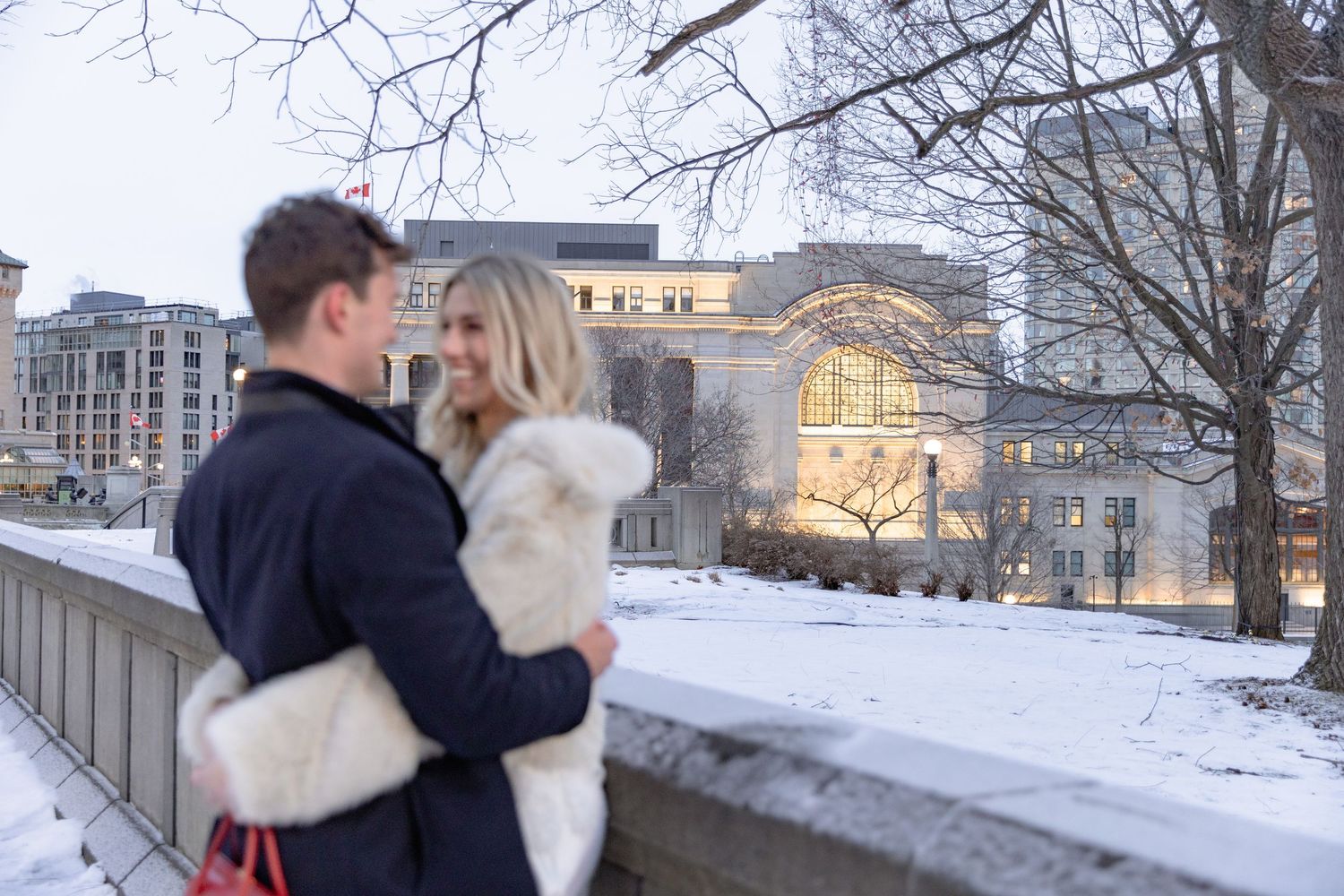 A couple embraces in a snowy winter scene with buildings and bare trees in the background.