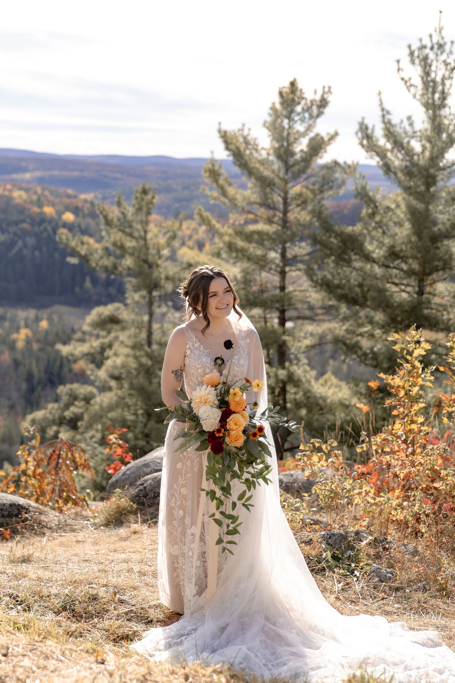A bride in a white lace gown poses on a mountaintop with evergreen trees and autumn foliage in the background.