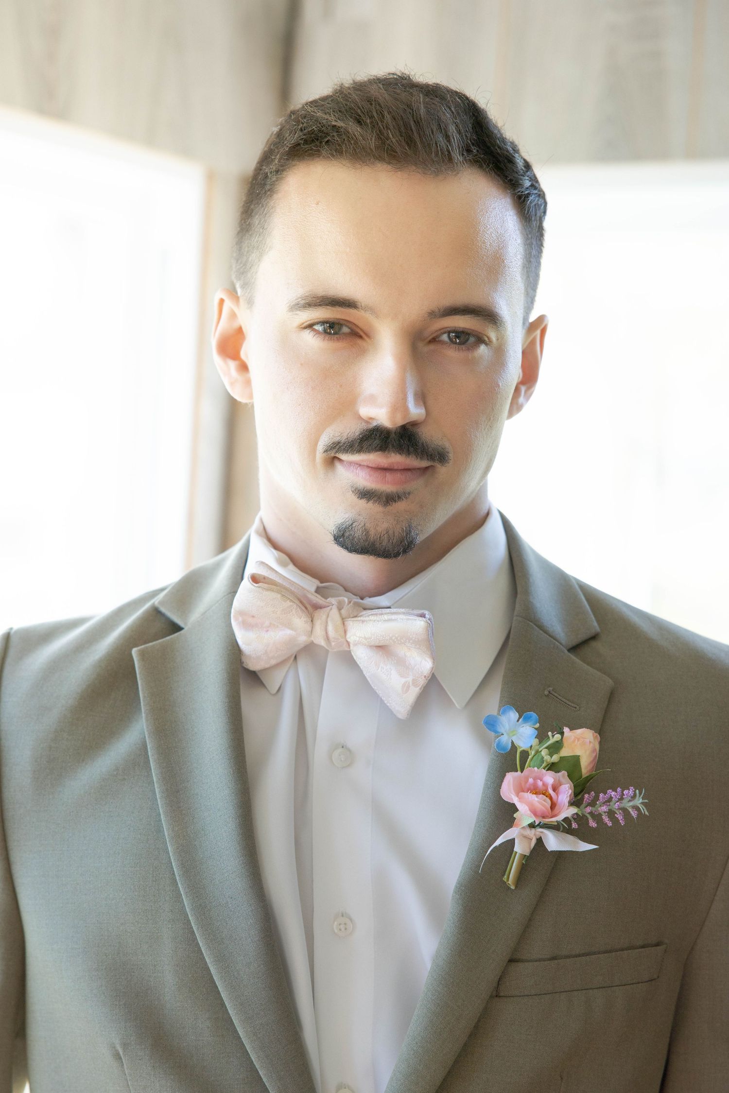 A groom in a gray suit and pink bow tie wears a delicate floral boutonniere with pink and blue blooms.