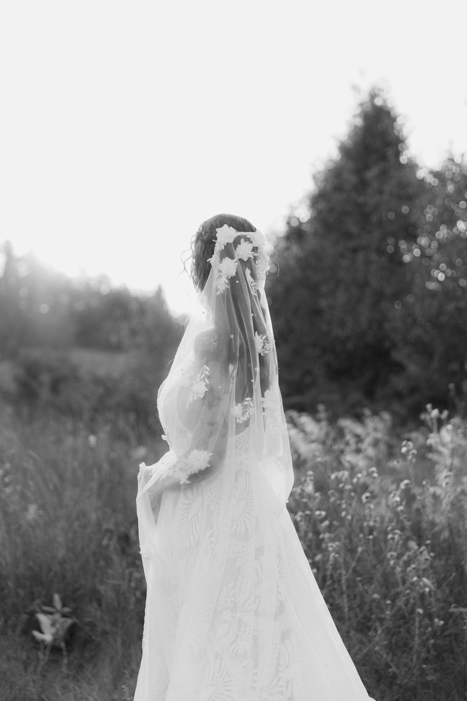 Black and white photo of a bride in a flowing lace wedding dress and veil standing in a sunlit meadow at dusk.