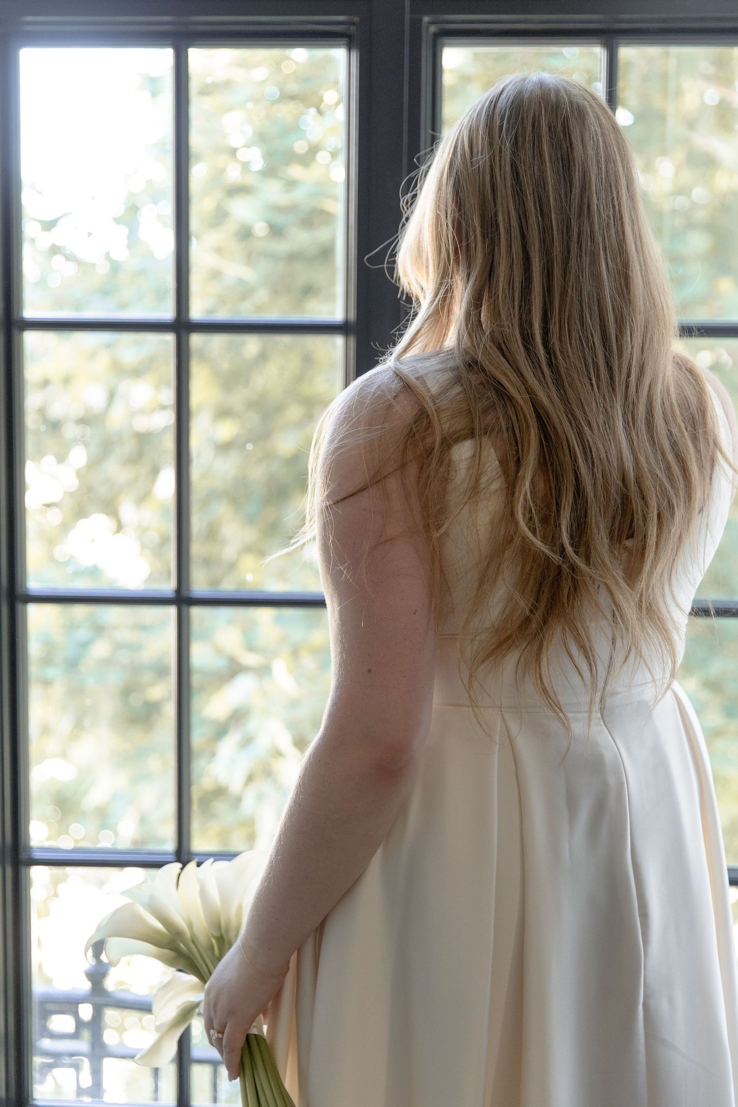 Person in white dress stands at large window holding flowers, photographed from behind with long blonde hair cascading down.