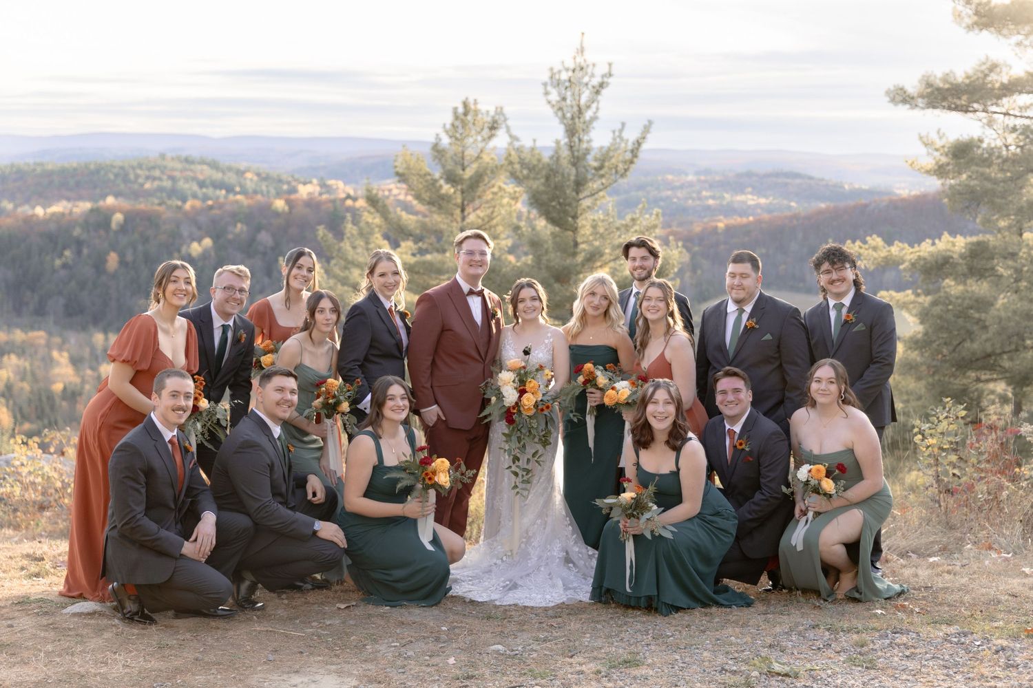Wedding party members sit together on a scenic mountain overlook at sunset.