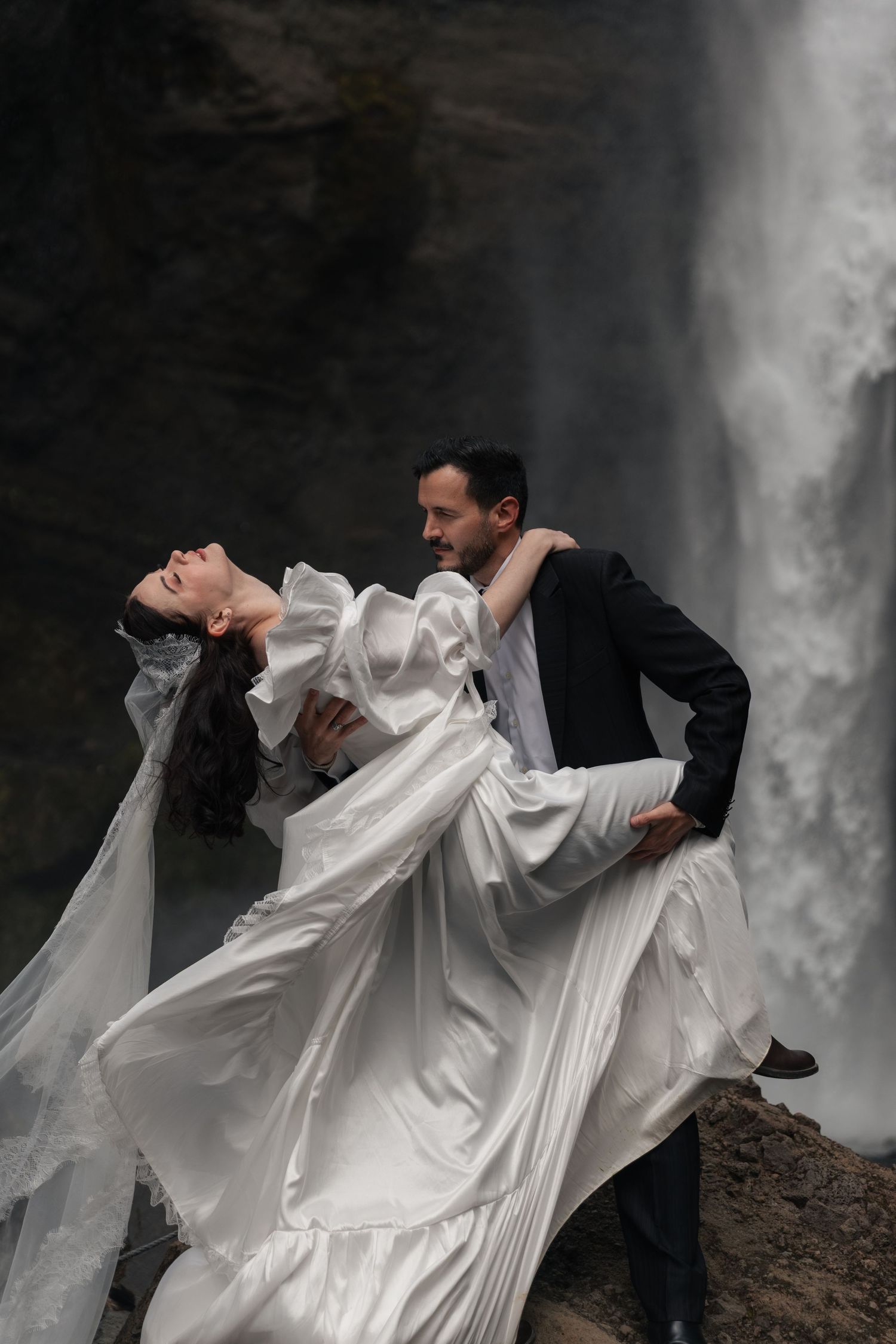 A groom dips his bride in a white flowing gown before a dramatic waterfall backdrop.