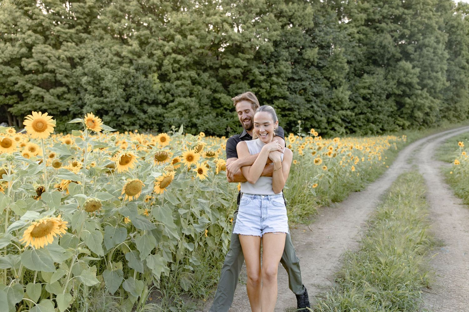 A couple stands on a dirt path next to a vibrant field of blooming sunflowers on a summer day.
