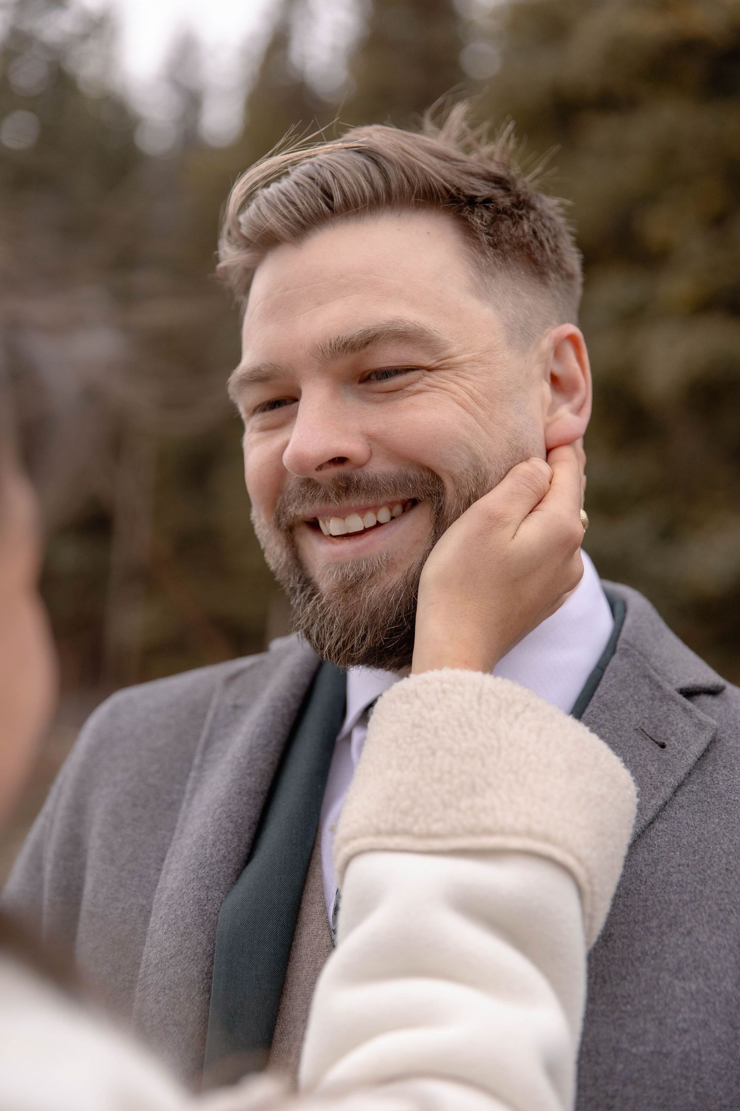 A person wearing a grey suit jacket smiles warmly while touching their face in a winter outdoor setting.