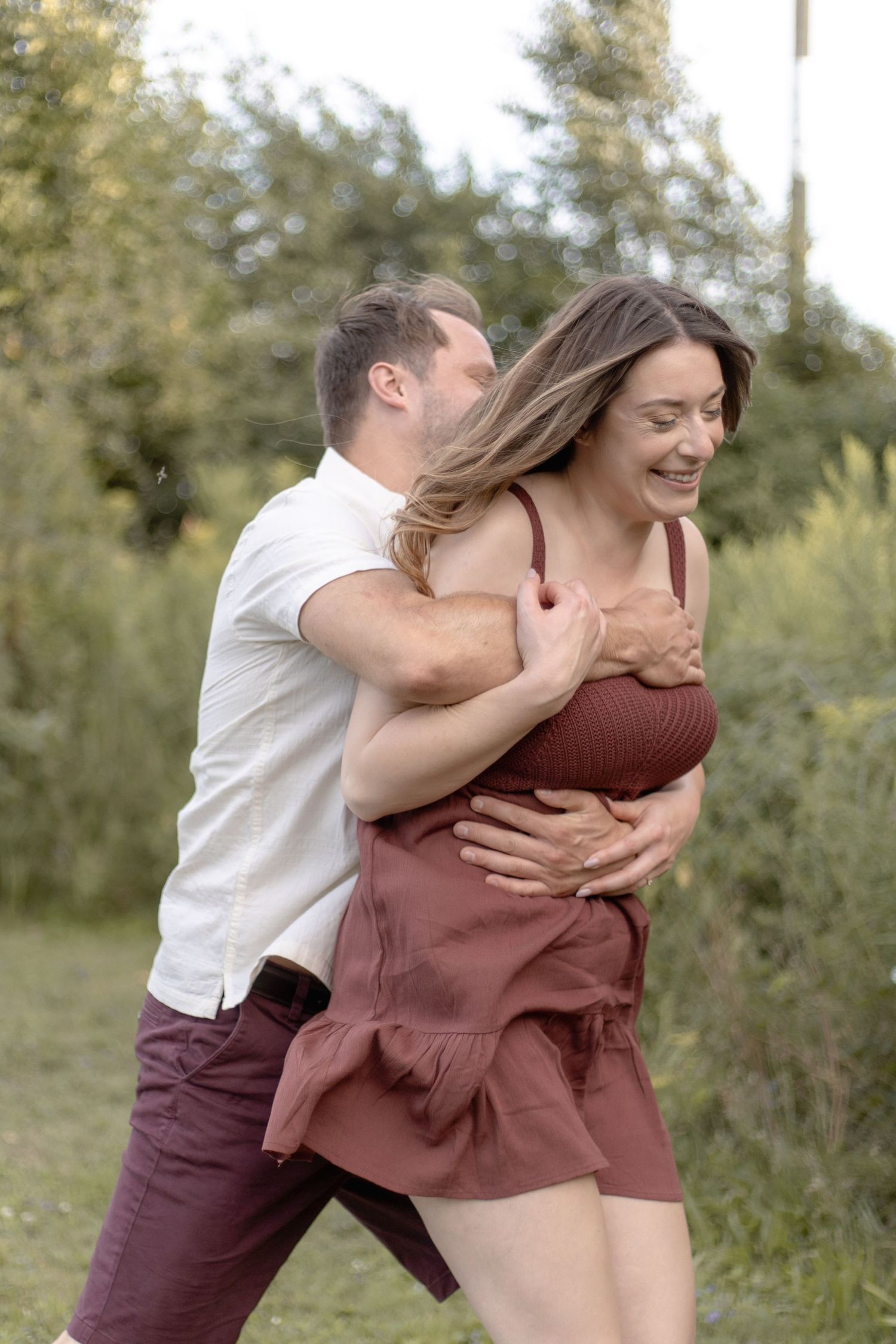 Two people share a playful embrace outdoors in a natural setting with trees and grass in the background.