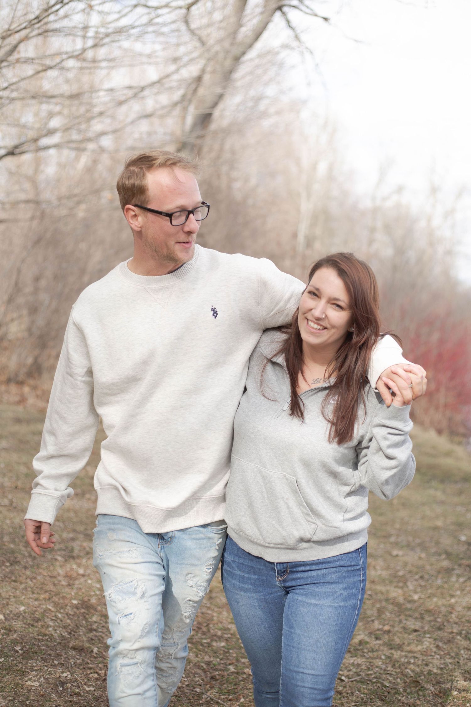 A couple wearing matching grey sweaters walks together on a foggy autumn path through bare trees.