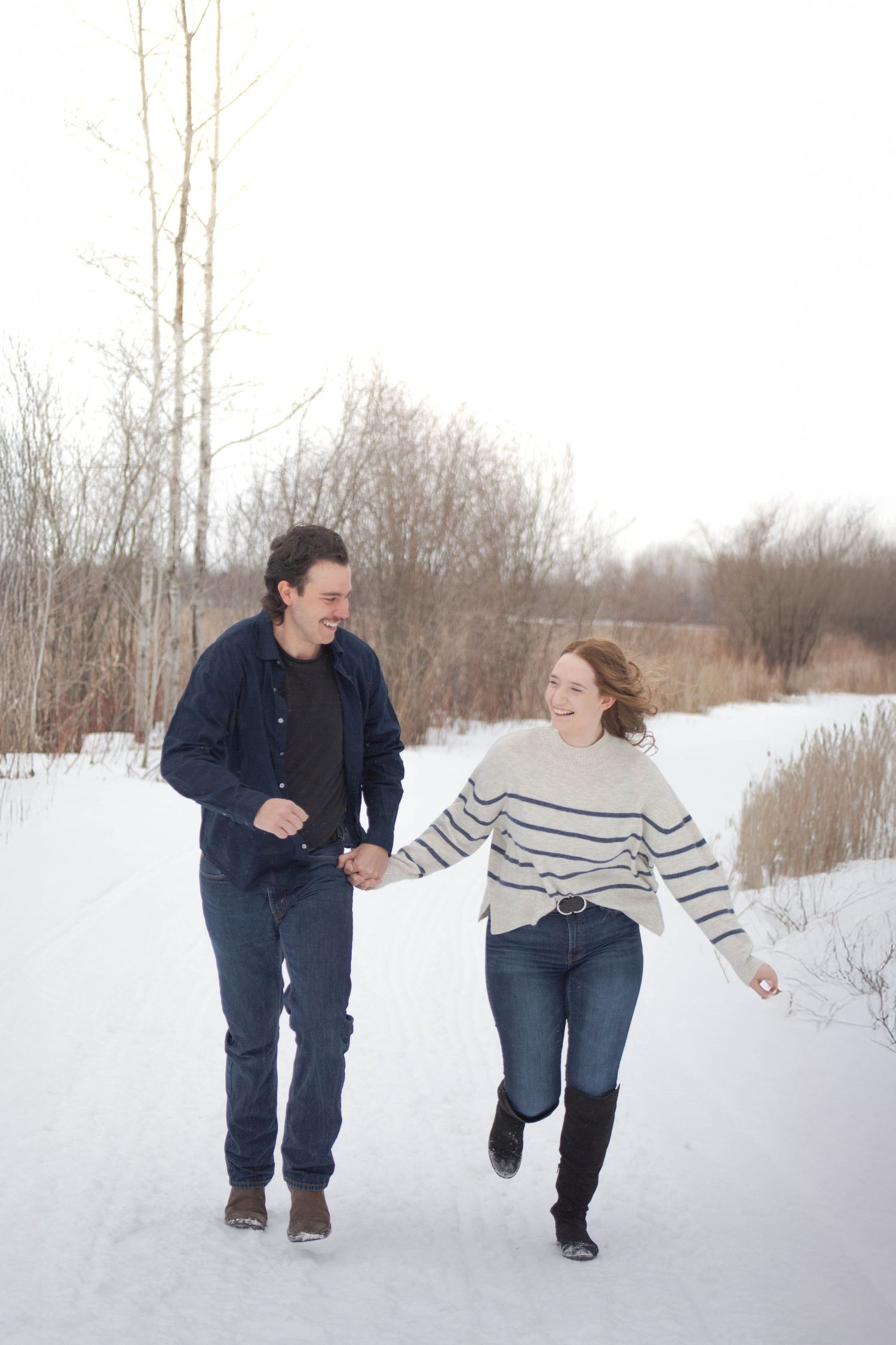 A couple runs playfully together on a snowy path surrounded by winter trees.