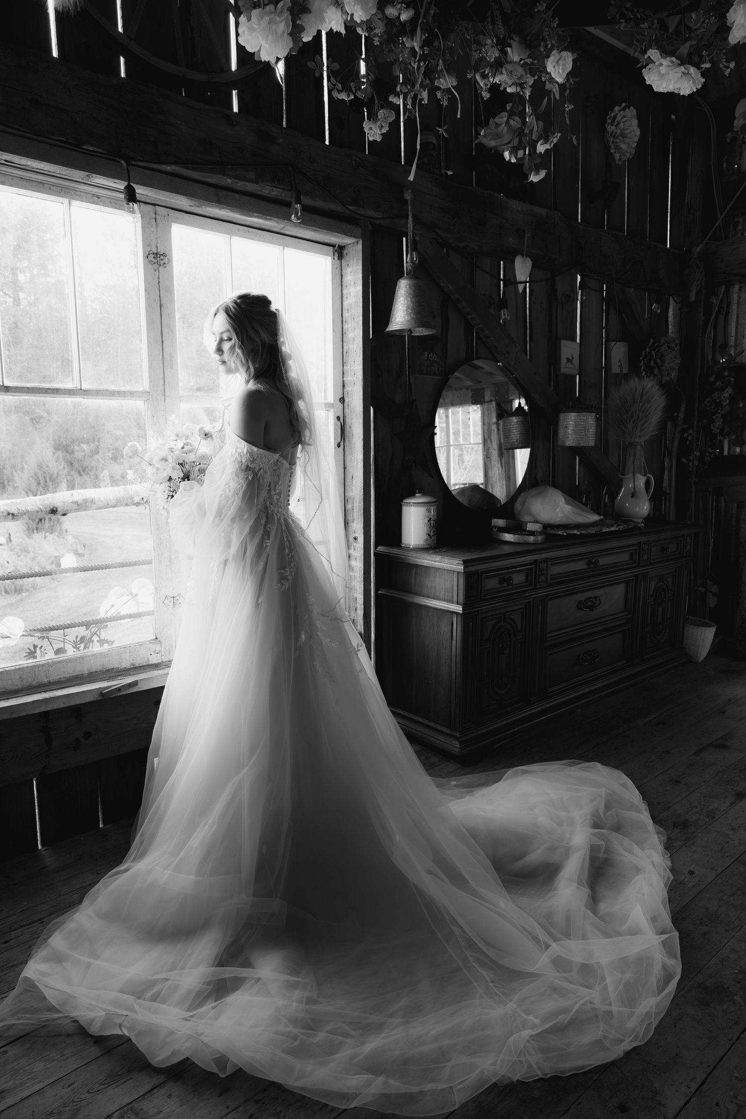 A dramatic black and white bridal portrait shows an ethereal silhouette standing by a window in a flowing tulle gown.