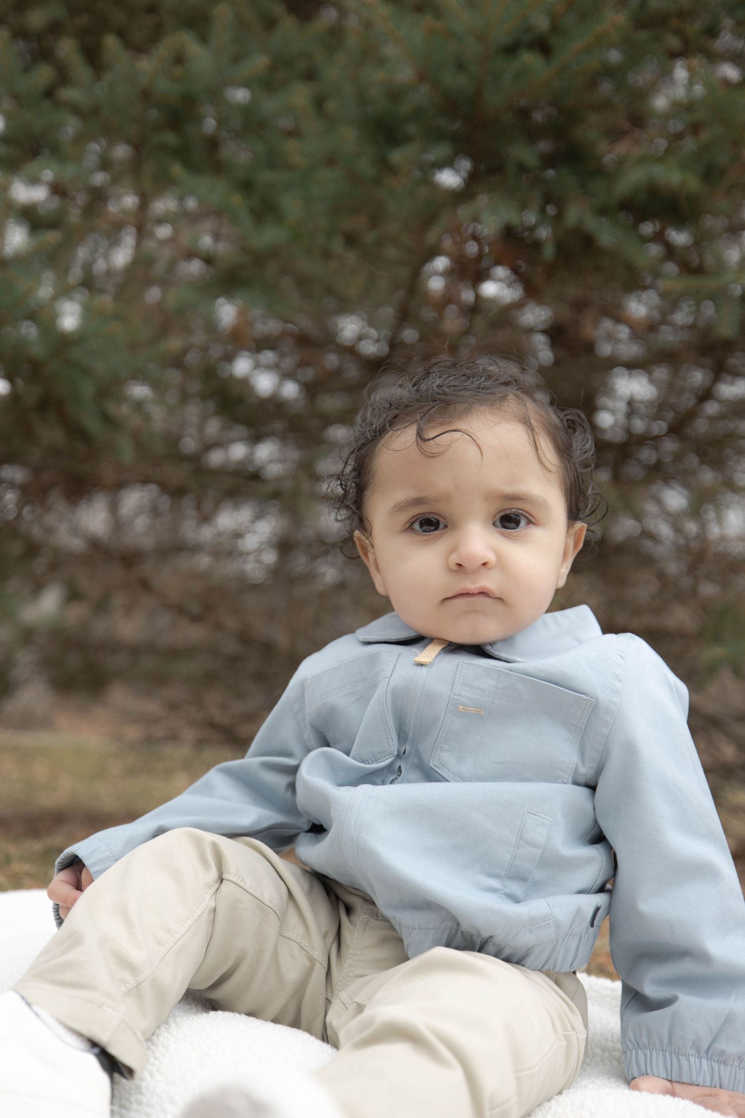 Young toddler in light blue jacket and khaki pants sitting outdoors against greenery background.