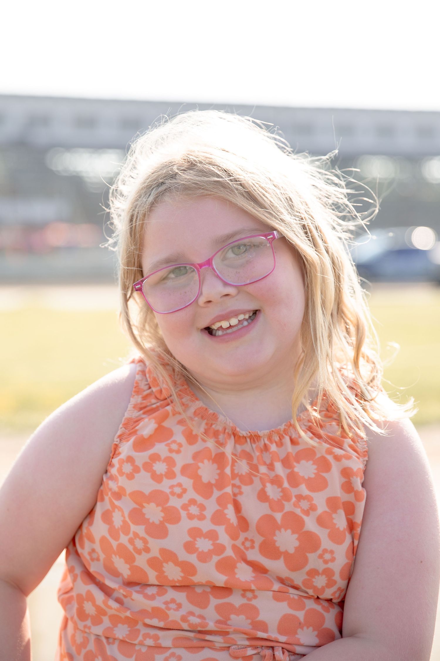 Young person wearing pink glasses and a sleeveless orange floral dress smiles brightly in outdoor sunlit setting.