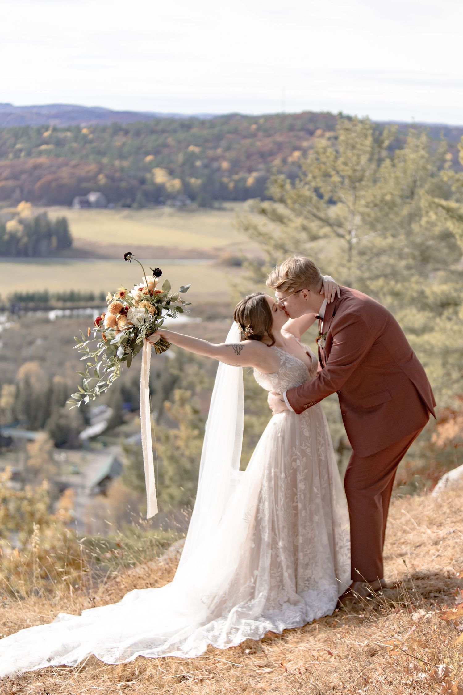 A couple in wedding attire sits on a cliff edge overlooking a scenic autumn mountain landscape at sunset.