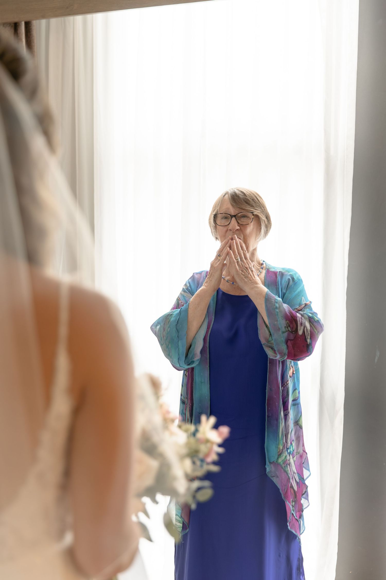 Person in blue dress and colorful shawl stands by window with hands clasped in emotional reaction.