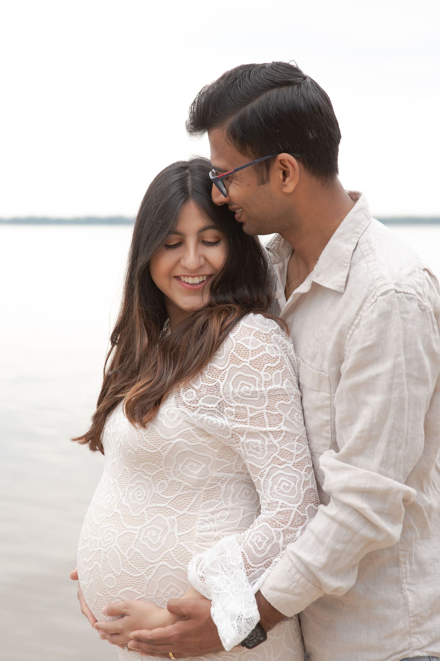 Romantic maternity photo of a couple in white outfits embracing by the water at sunset.