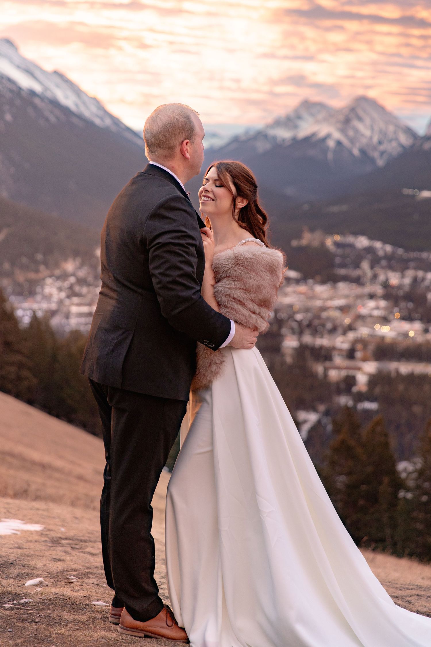 A couple shares a romantic moment at sunset in front of snow-capped mountains, the bride wearing a white gown and fur wrap.