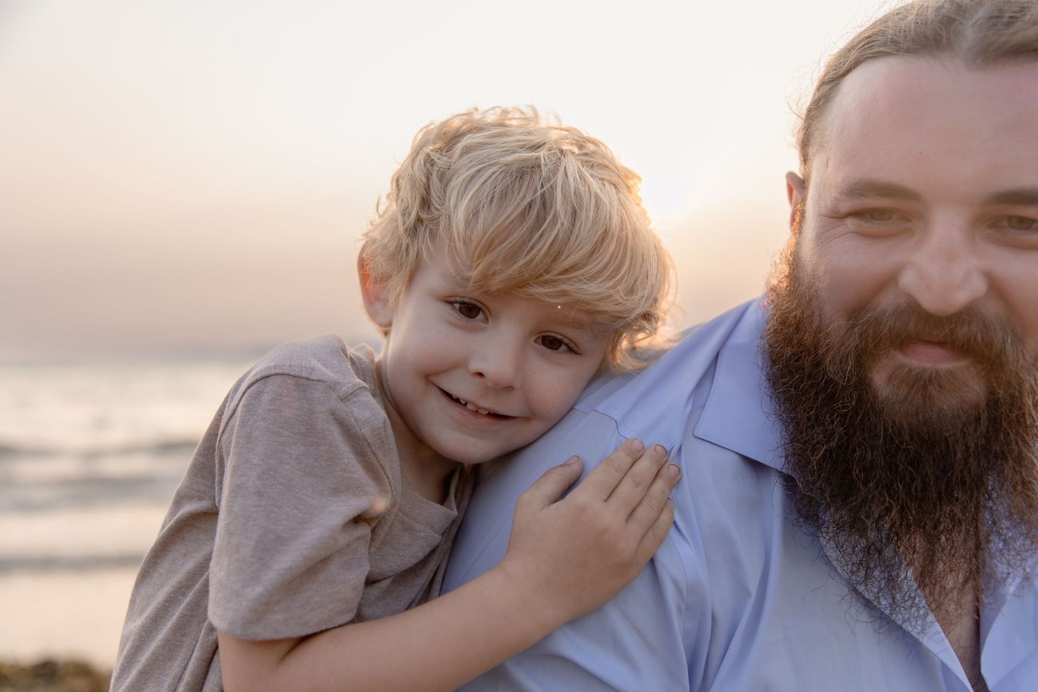 A sweet moment captured at sunset with a bearded adult and small child sharing an embrace on the beach.
