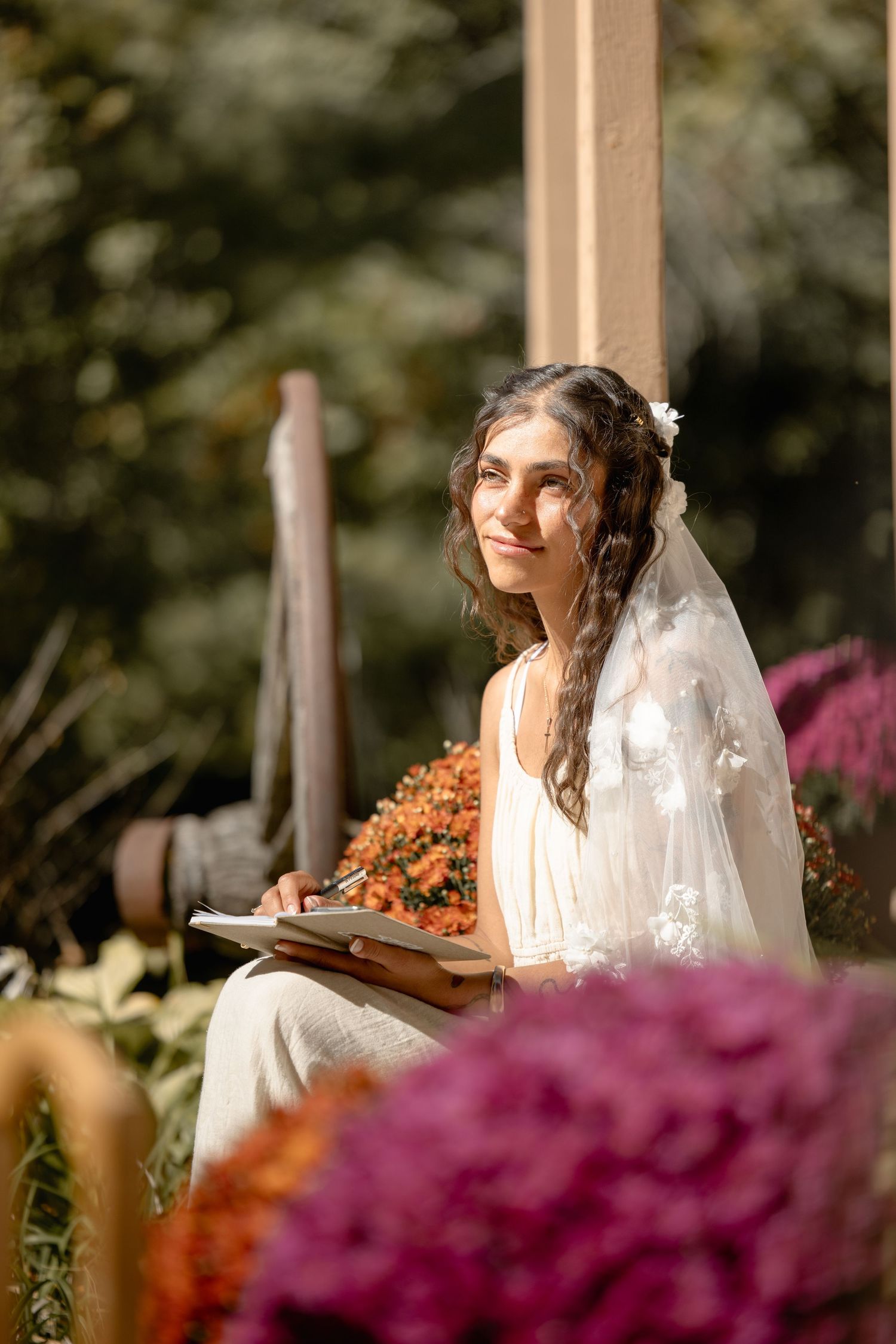 A bride in a white wedding dress sits among vibrant purple and orange flowers in a garden setting.
