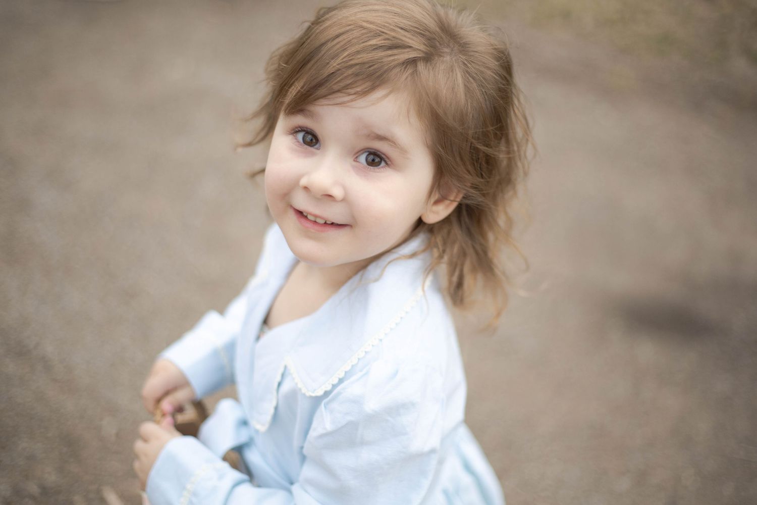Young child wearing a light blue and white dress smiles while looking up at the camera during an outdoor portrait session.