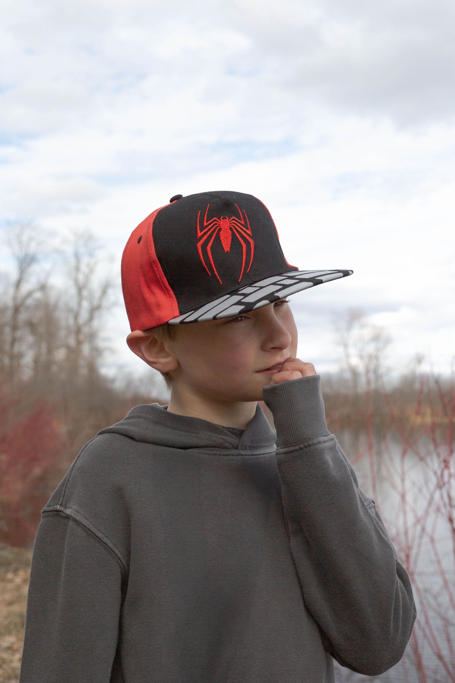 Young boy wearing a Spider-Man baseball cap and grey hoodie gazes thoughtfully into the distance.