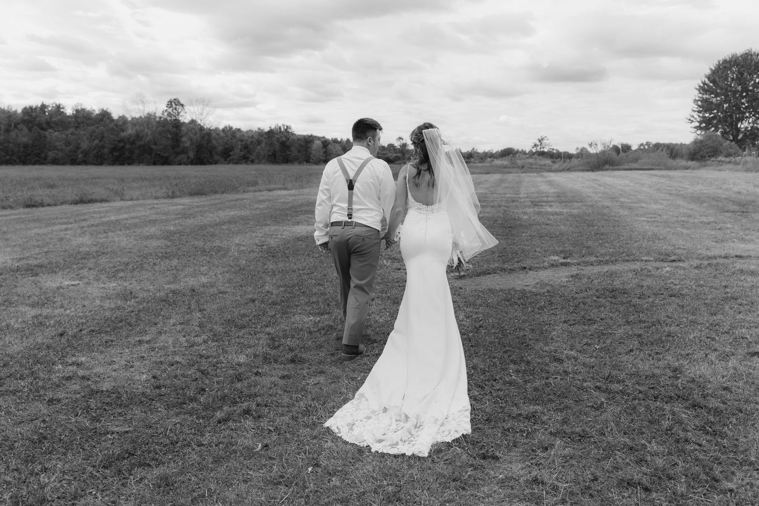 A couple in wedding attire walks together across a grassy field under cloudy skies.