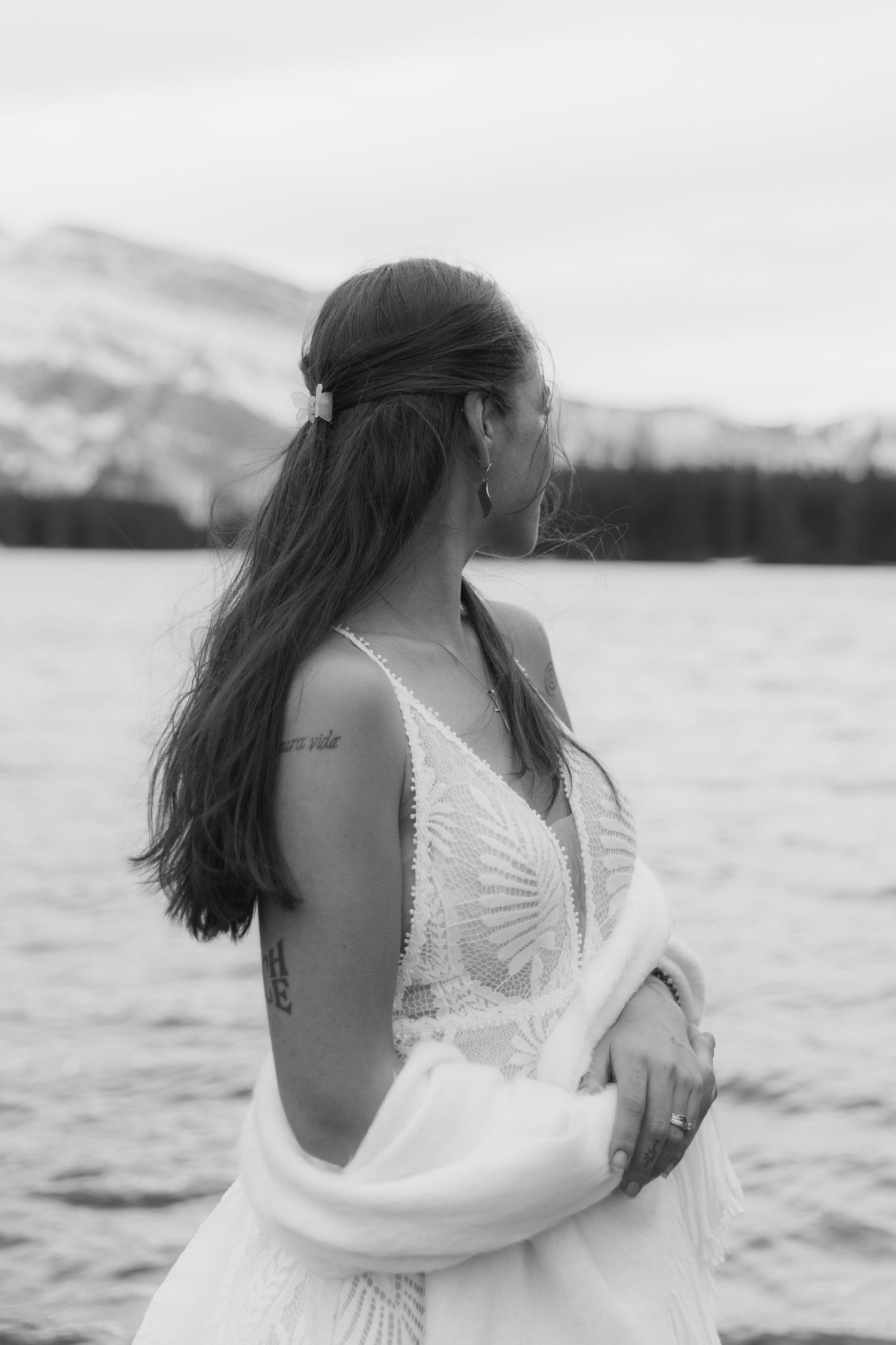 A couple embraces in white attire by a scenic mountain lake in a black and white romantic photograph.