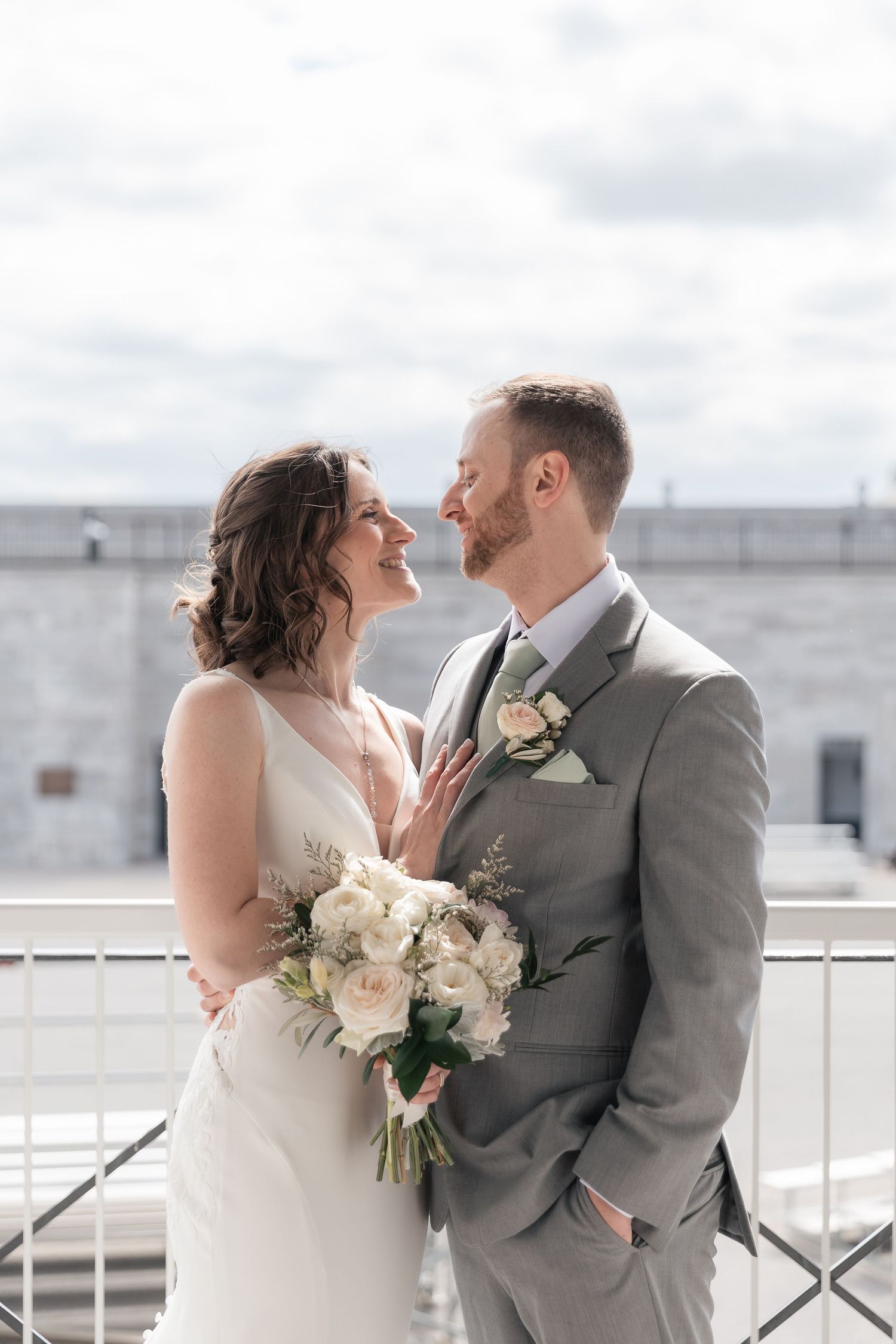 Smiling bride and groom in gray suit share a tender moment on a rooftop balcony, bride holding white roses.