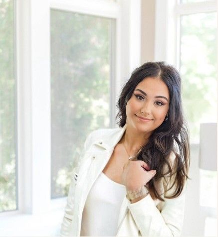 Woman with dark hair wearing a white sweater poses in front of bright window light.