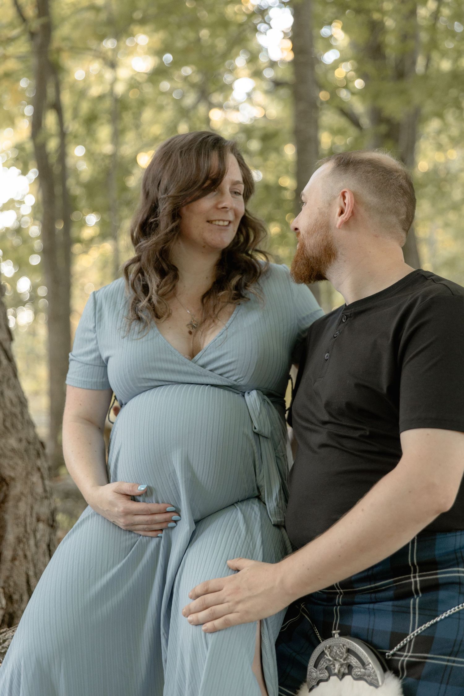 A couple shares an intimate moment in the woods, with the person in a flowing blue maternity dress and their partner in a kilt.