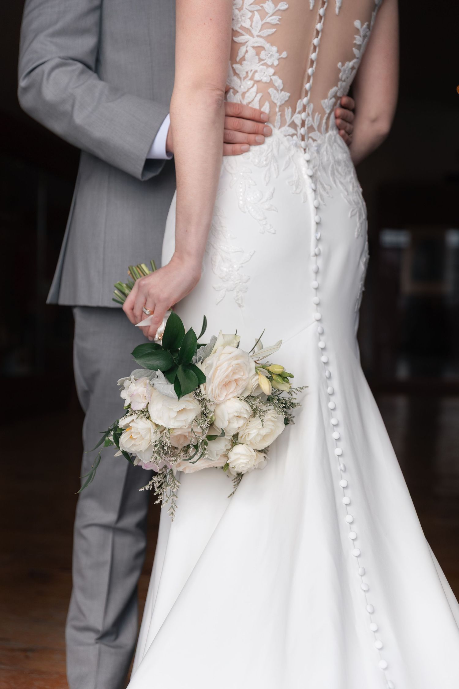 Bride in lace back wedding gown holding white rose bouquet with groom in gray suit at wedding ceremony.