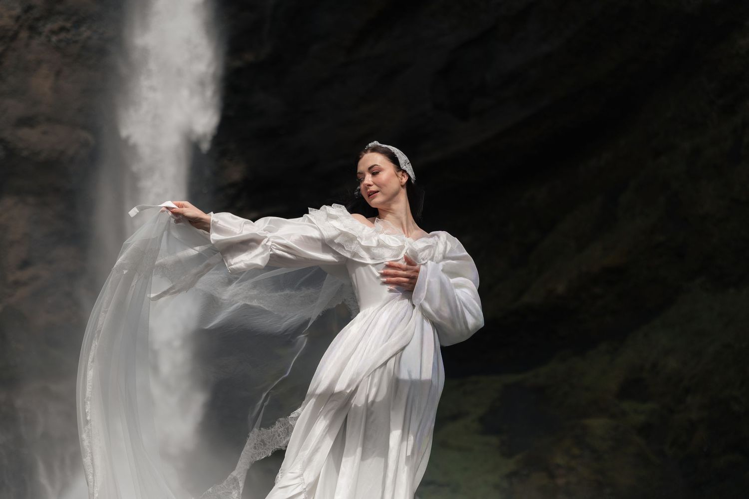 Elegant bride in white gown with billowing sleeves poses gracefully in front of a majestic waterfall.