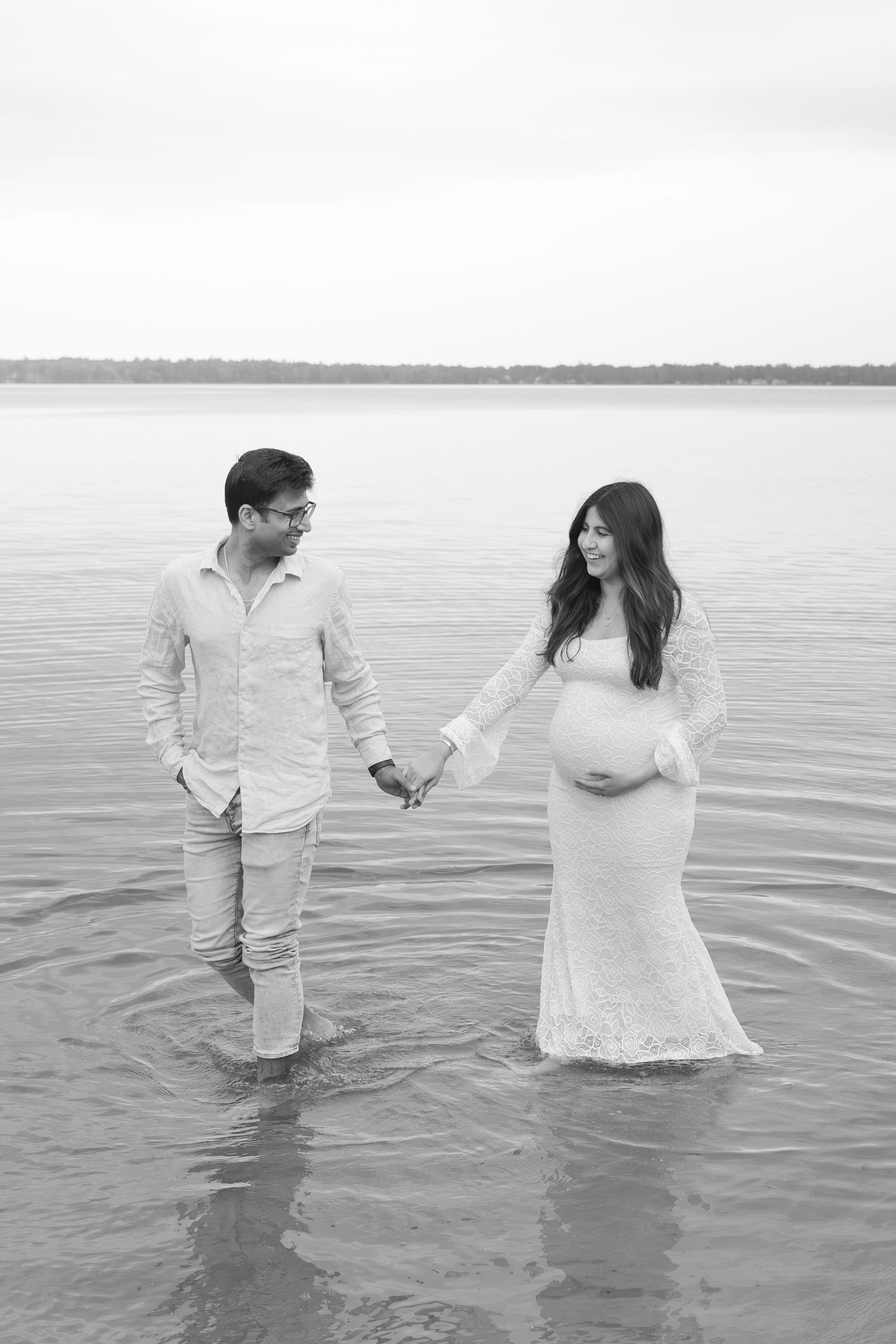 A couple stands together in shallow lake water with light ripples around them, captured in black and white.