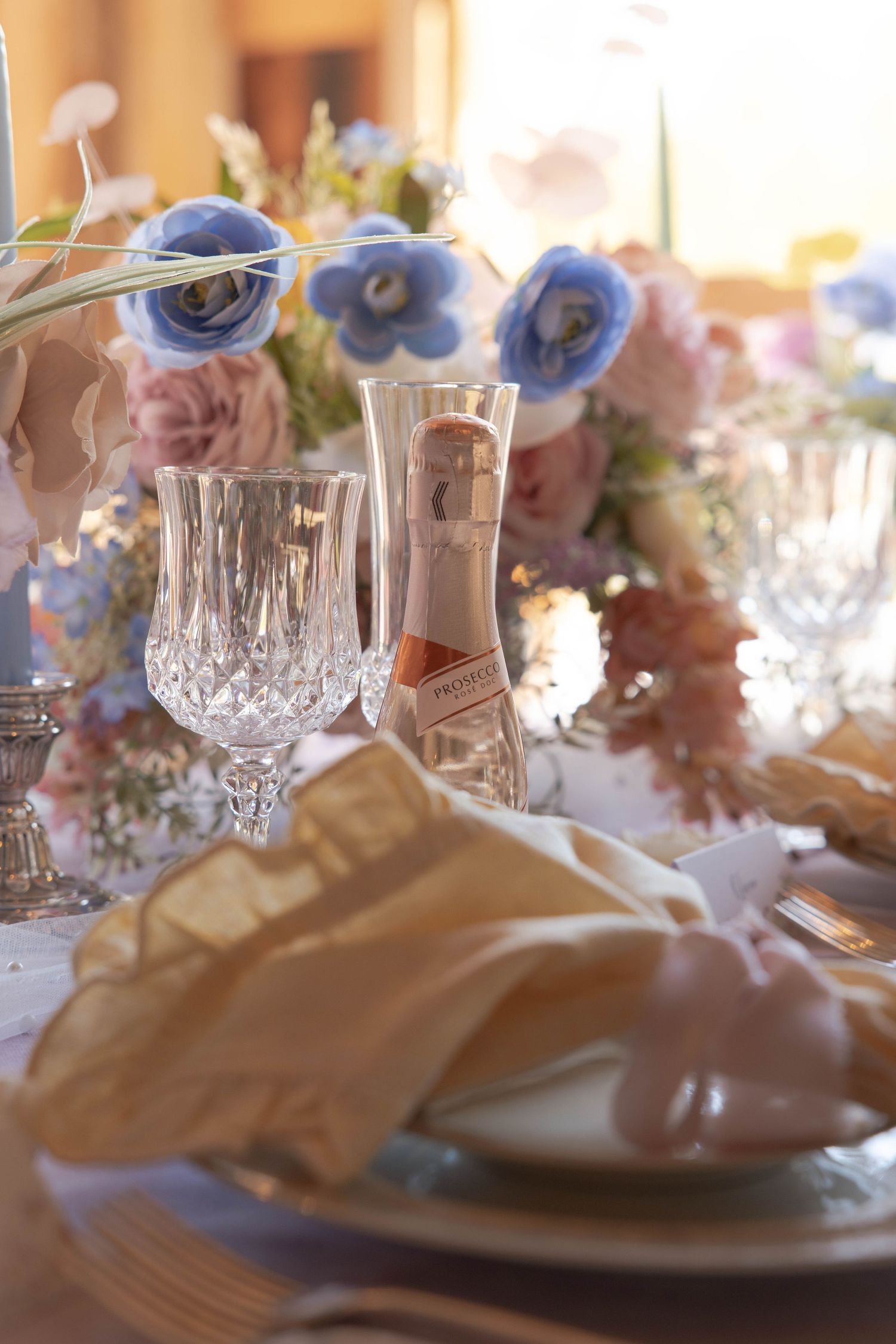 Elegant table setting with crystal glassware, blue and pink flowers, and soft fabric napkins in warm lighting.