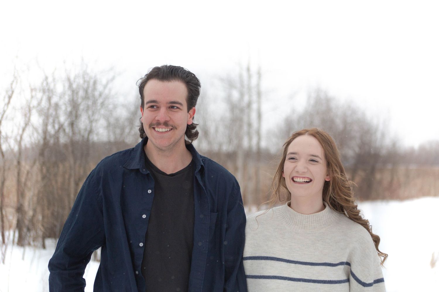 A cheerful winter portrait of a couple standing in the snow with bare trees in the background.