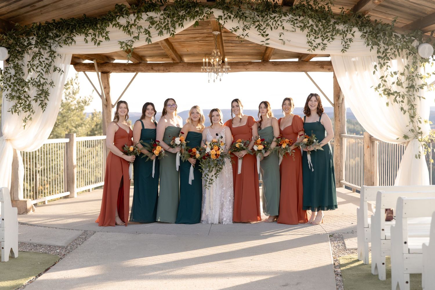 A wedding party in colorful dresses poses together in a scenic outdoor setting during golden hour.