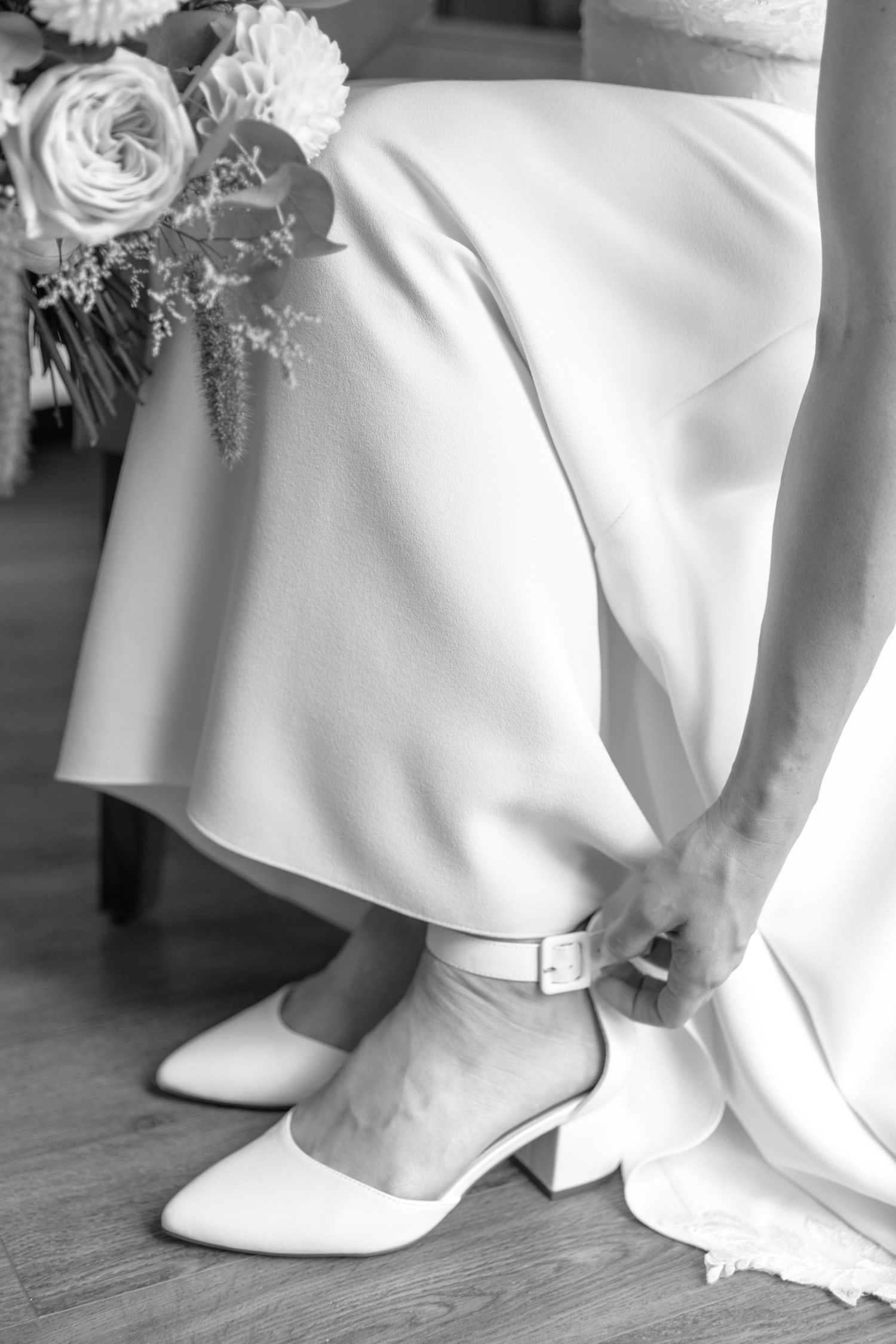 Row of elegant white wedding shoes peeking out from beneath flowing bridal gowns on a wooden floor.