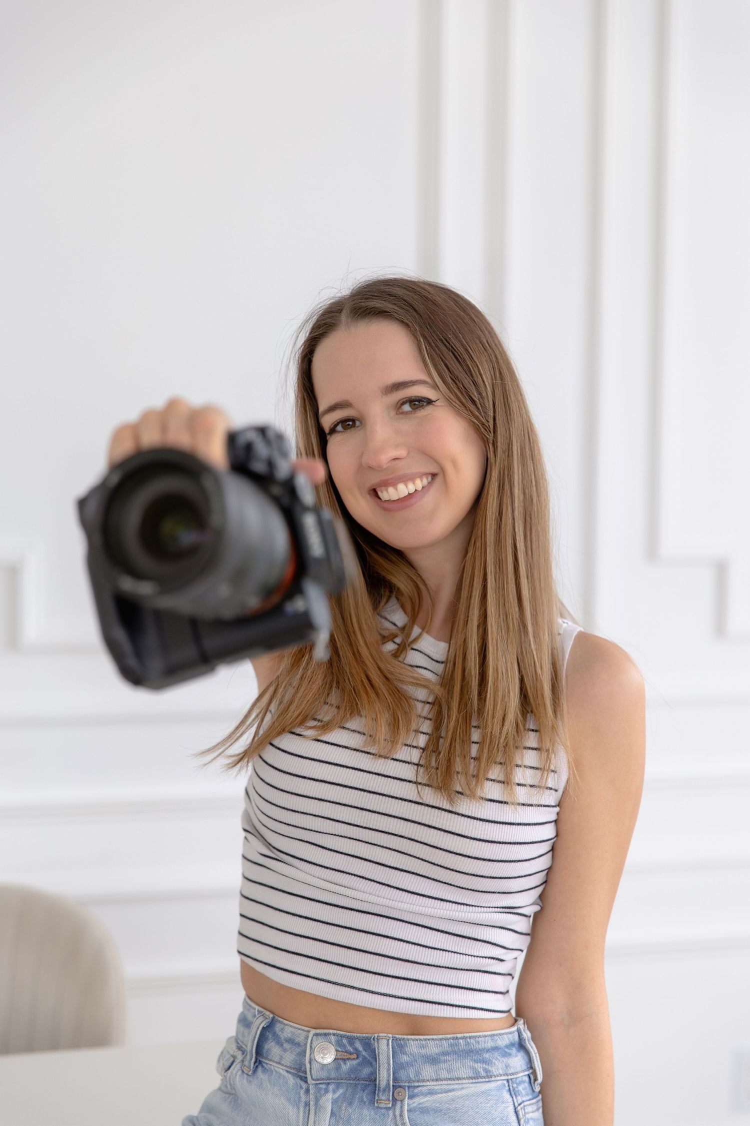 A photographer in a striped crop top and jeans poses with a DSLR camera in a bright white studio setting.