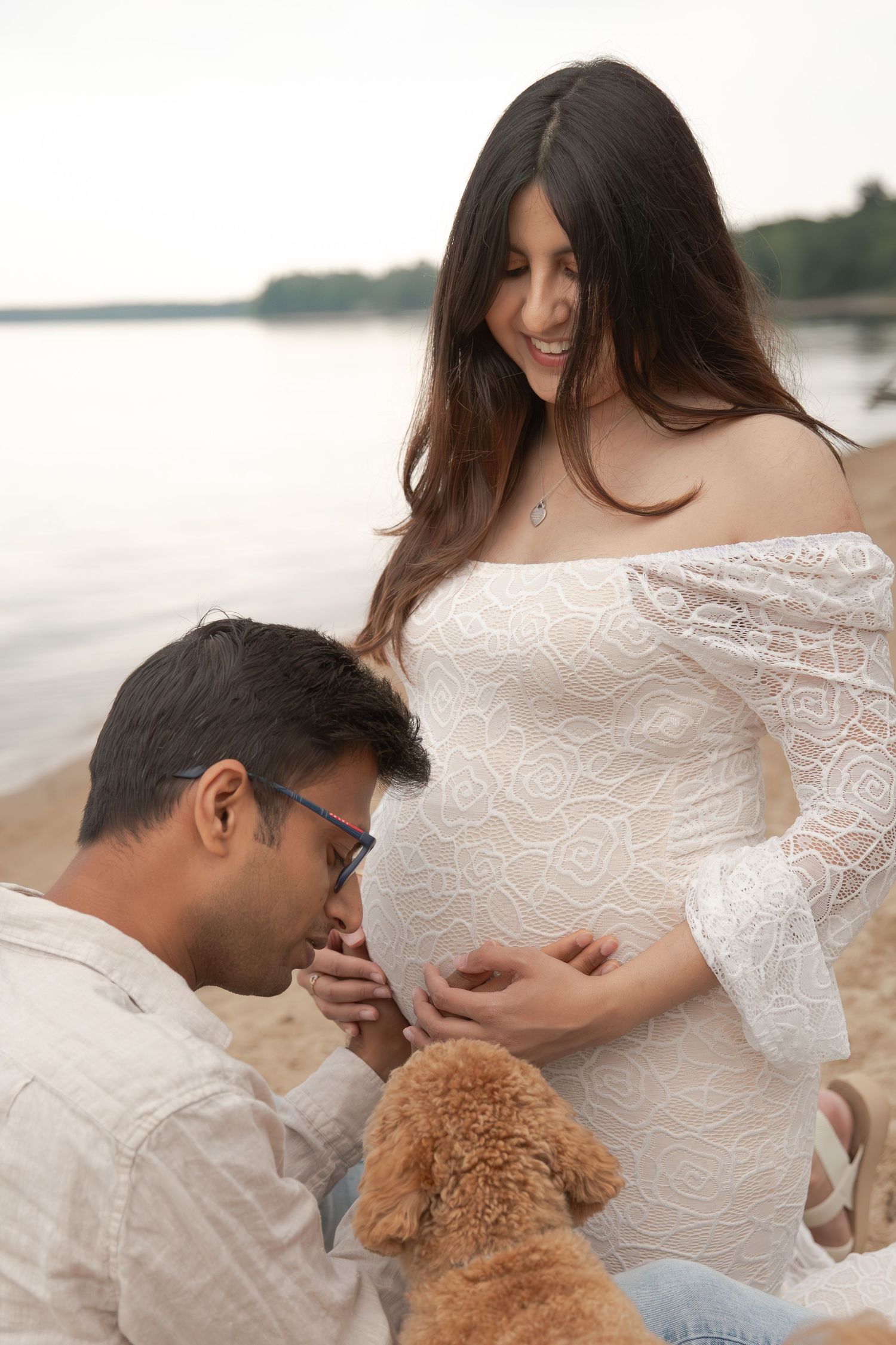 A couple shares an intimate moment on the beach with their dog while celebrating a pregnancy in a lace white dress.