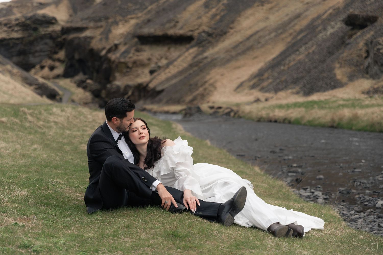 Bride and groom sitting together on grass by a river with dramatic rocky cliffs in Iceland wedding photo.