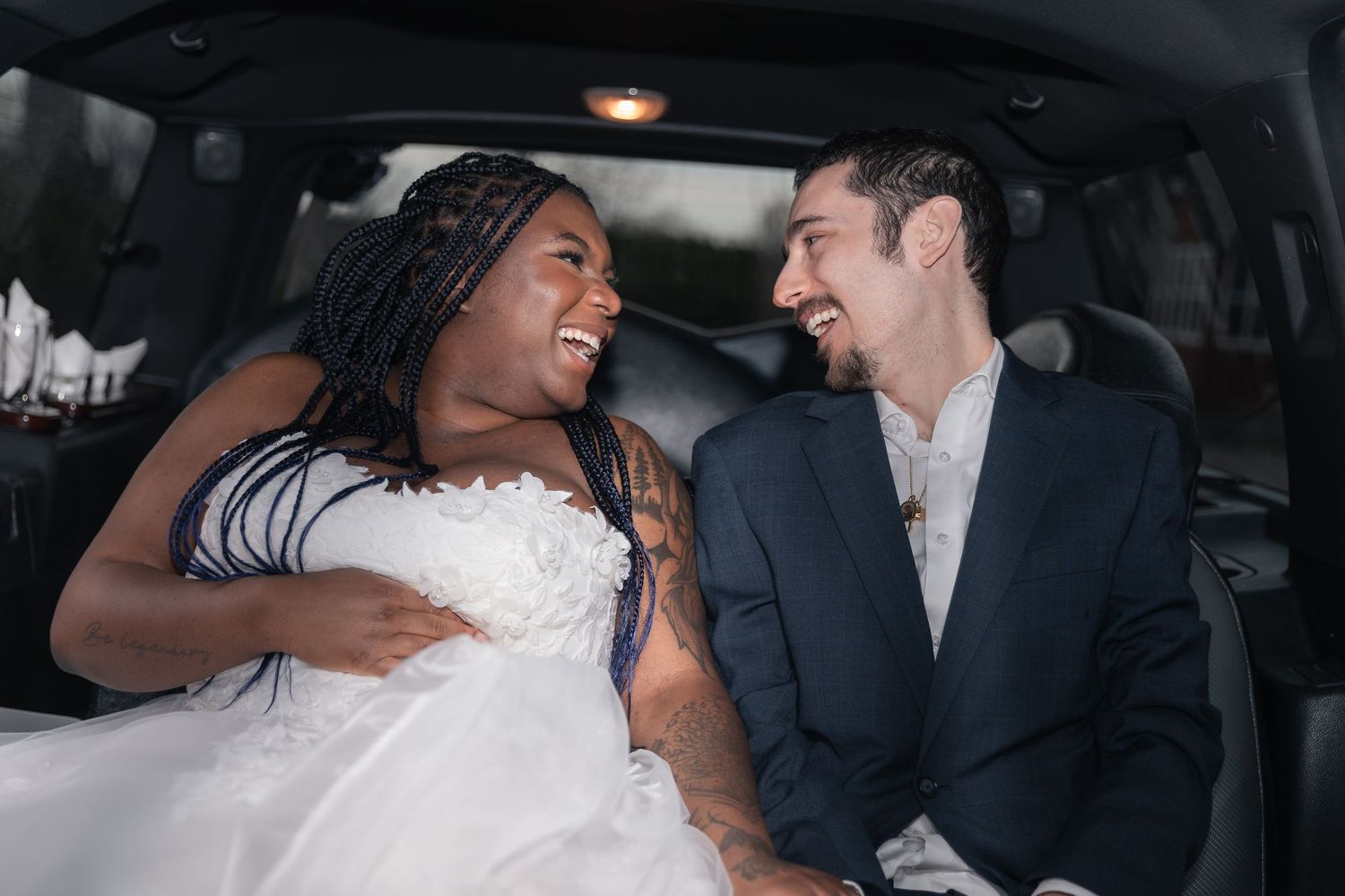 Happy interracial newlywed couple laughing together in a limousine, bride in white gown and groom in navy suit.