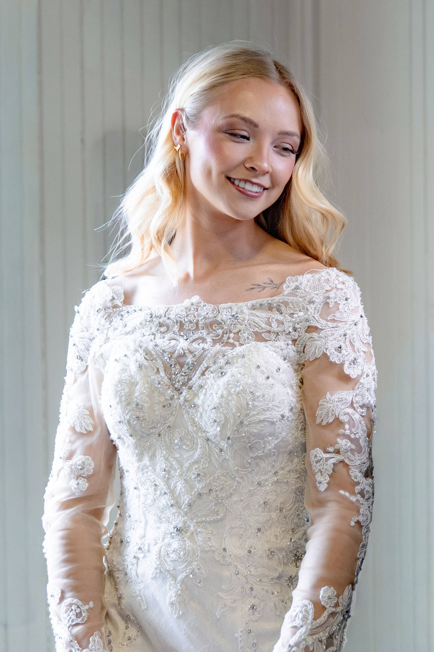 A bride in a long-sleeved lace wedding dress with intricate beading and embellishments poses in a light-filled room.