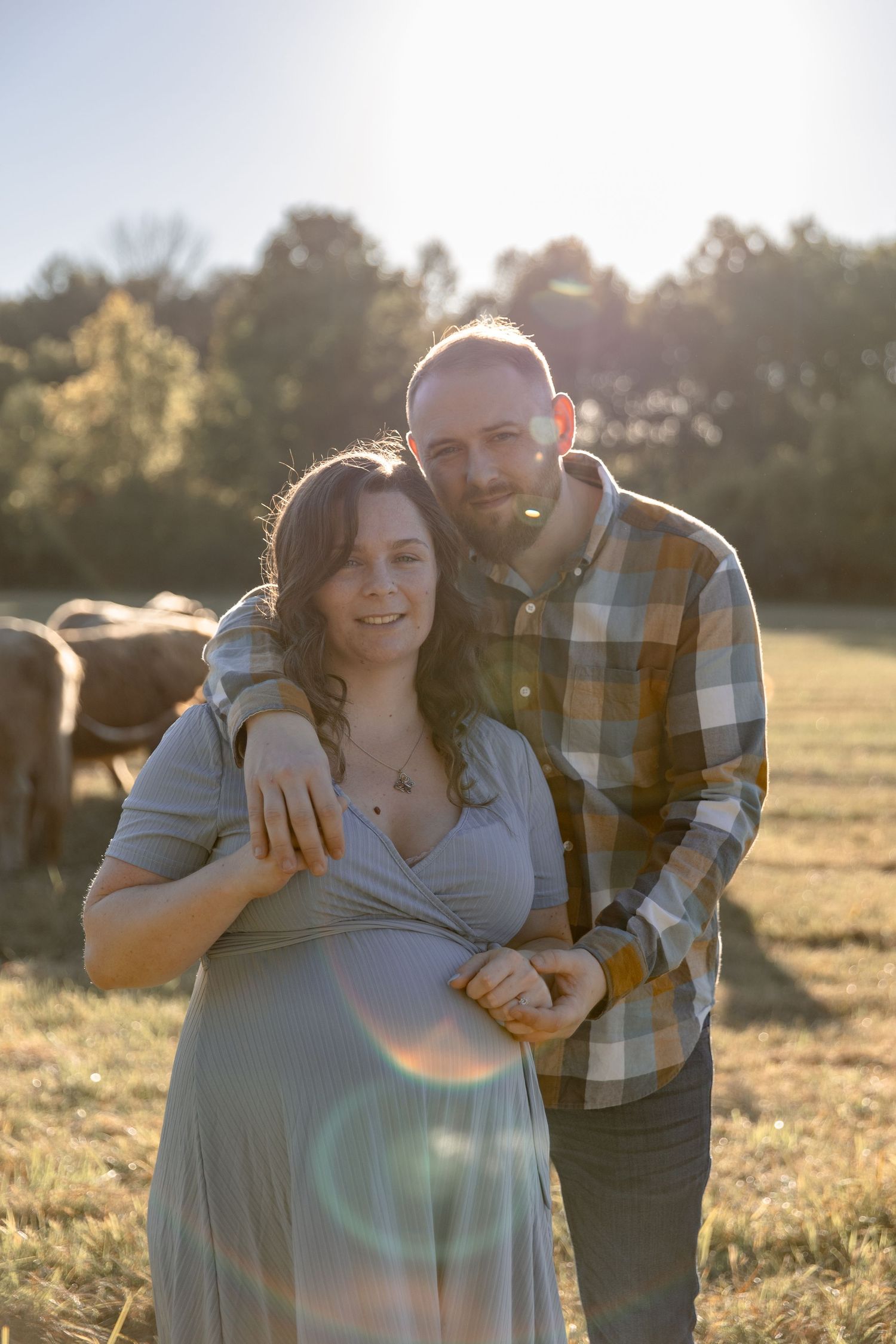 A couple embraces in a sunlit field during golden hour with lens flare and trees in the background.