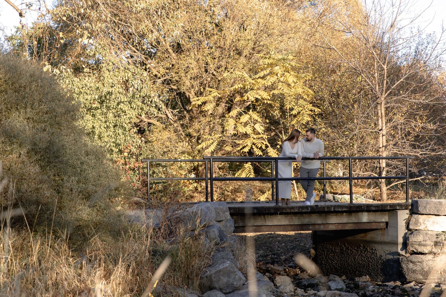 A person walks across a wooden footbridge surrounded by autumn trees and foliage in a peaceful park setting.