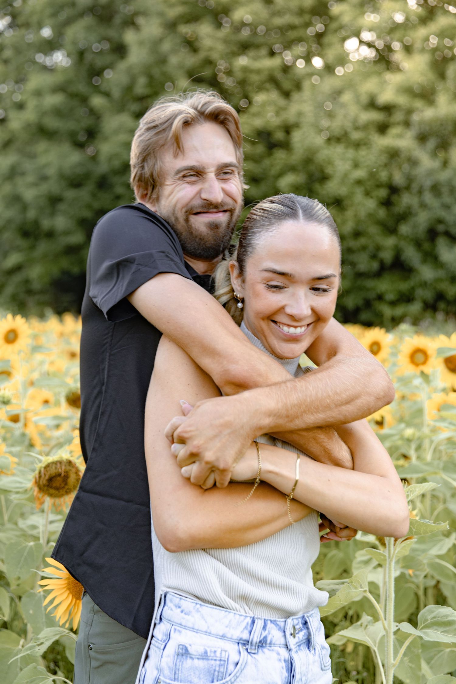 A couple embraces playfully in a sunlit sunflower field during a warm summer day.
