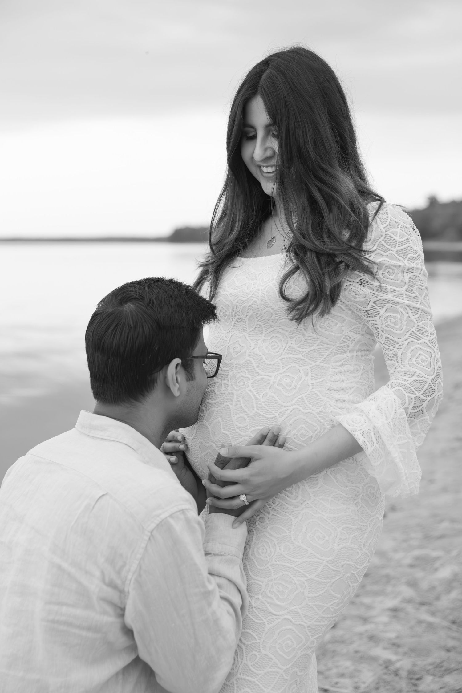 Black and white beach maternity photo of couple in white outfits sharing intimate moment along shoreline.