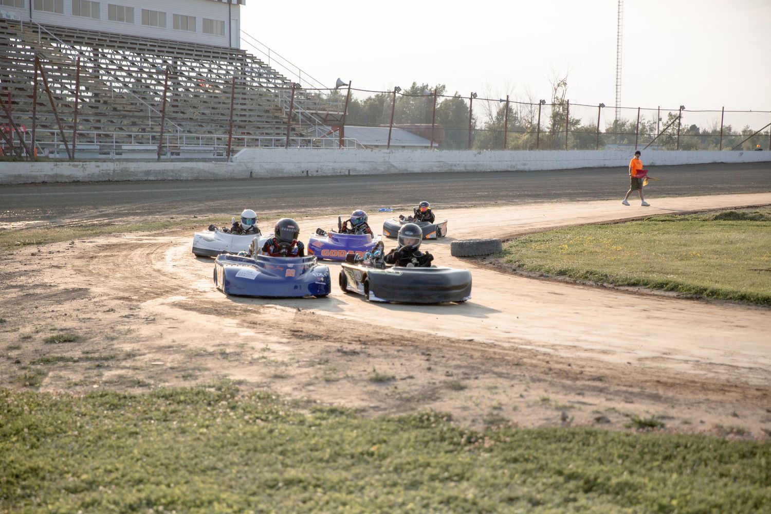 Go kart racers drive blue and purple bumper cars around a dirt track at a local racing venue.
