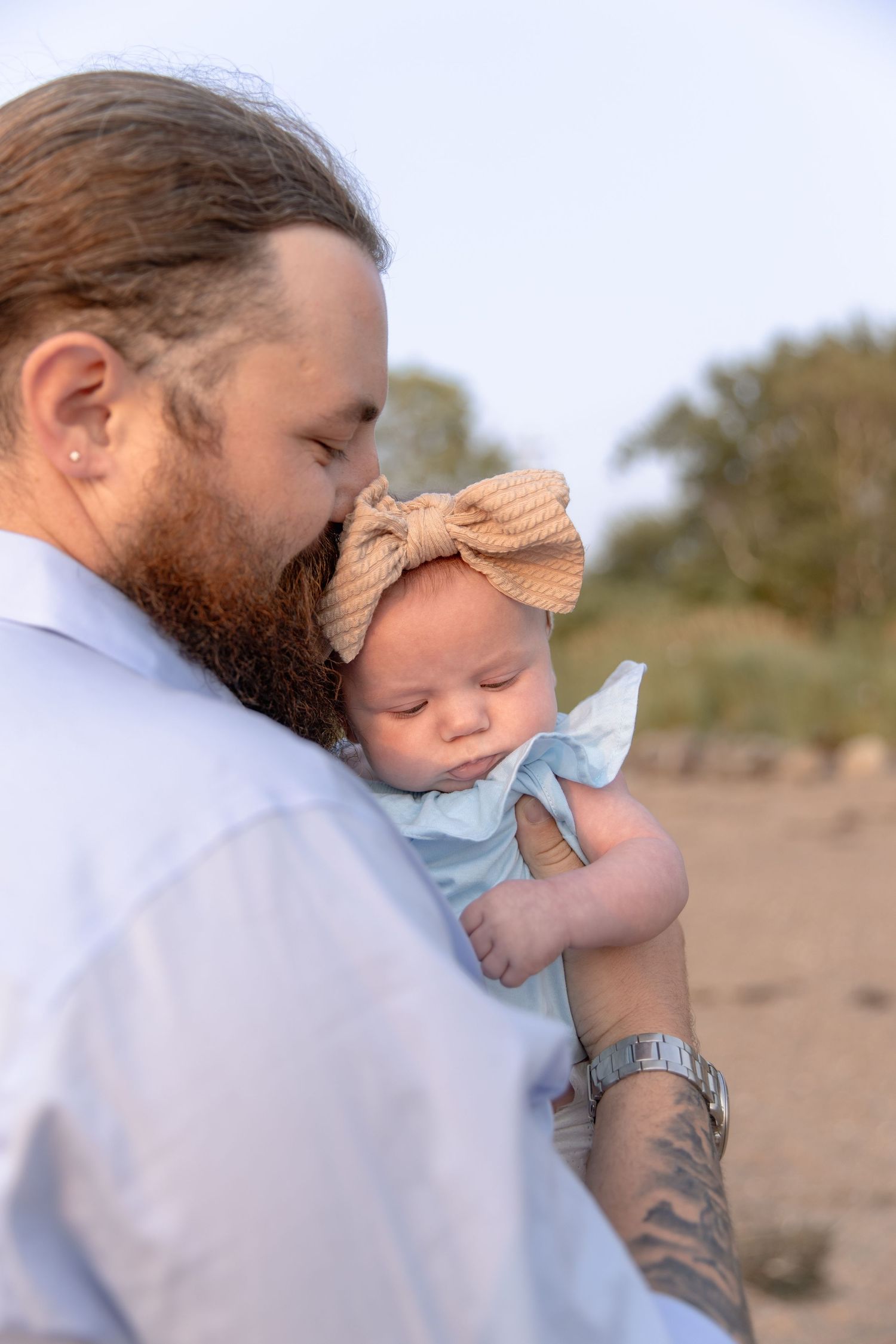 A bearded person wearing a blue shirt holds a sleeping baby with a bow headband outdoors.