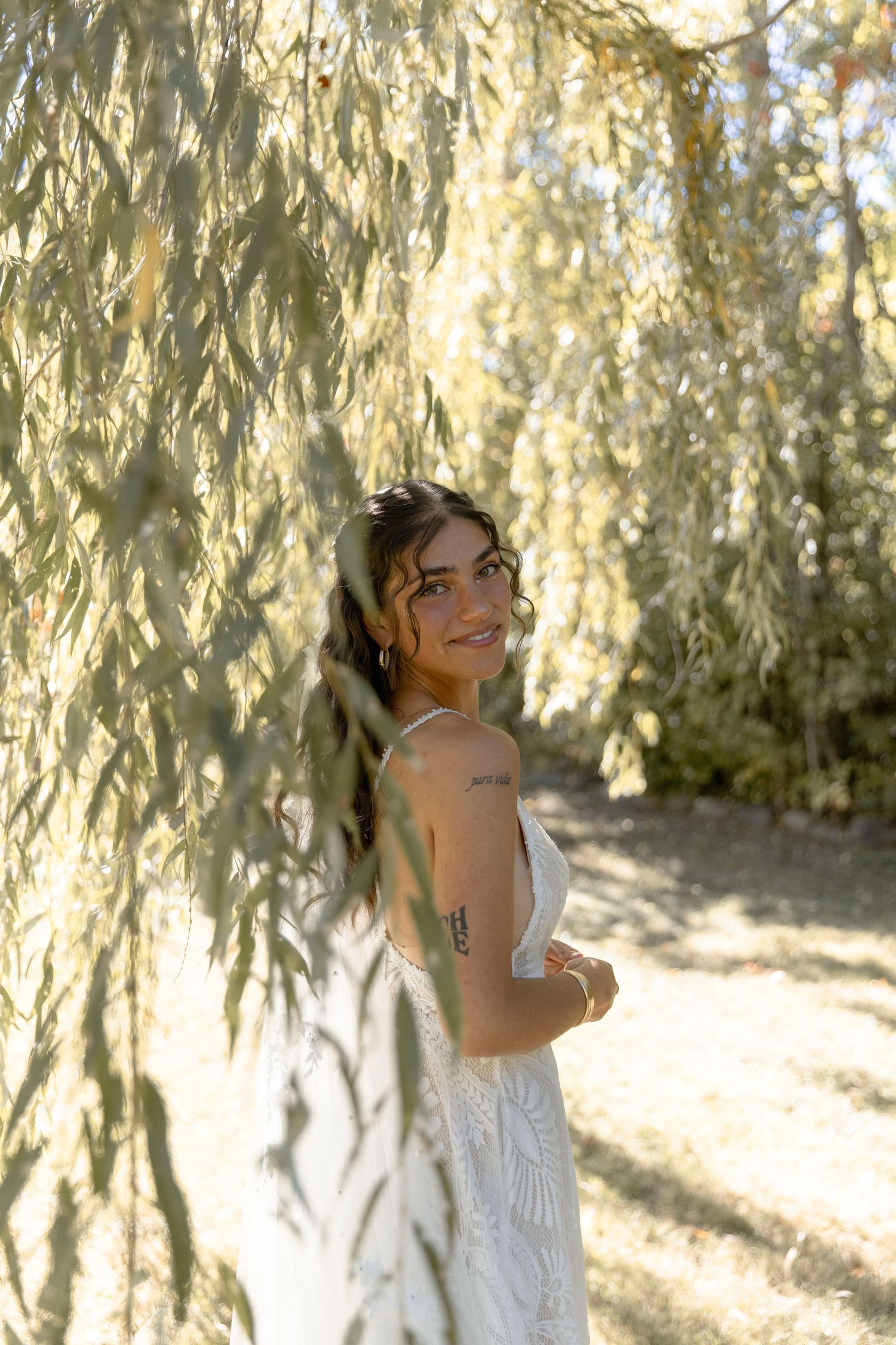 A person in a white dress dances joyfully under the cascading branches of a willow tree on a sunny day.